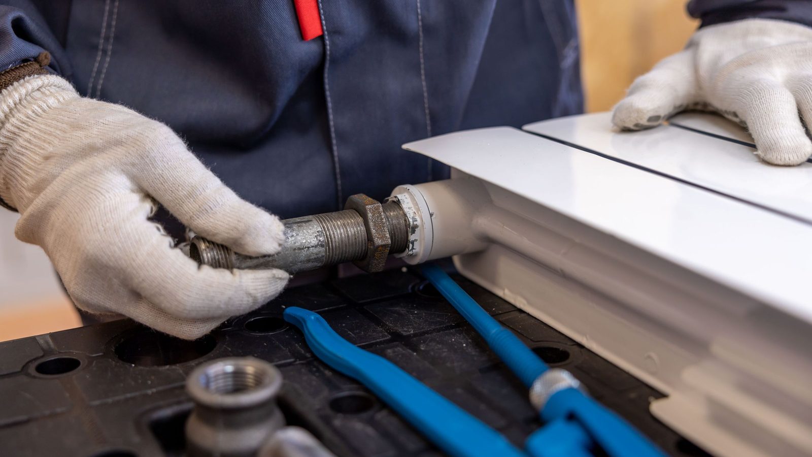 Plumber in work gloves fitting a threaded steel pipe, with blue pipe wrenches on the bench